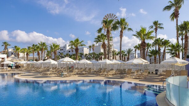 A pool area with sun loungers and umbrellas, surrounded by palm trees and a windmill. - Papantonia Hotel Apartments  (Paralimni)