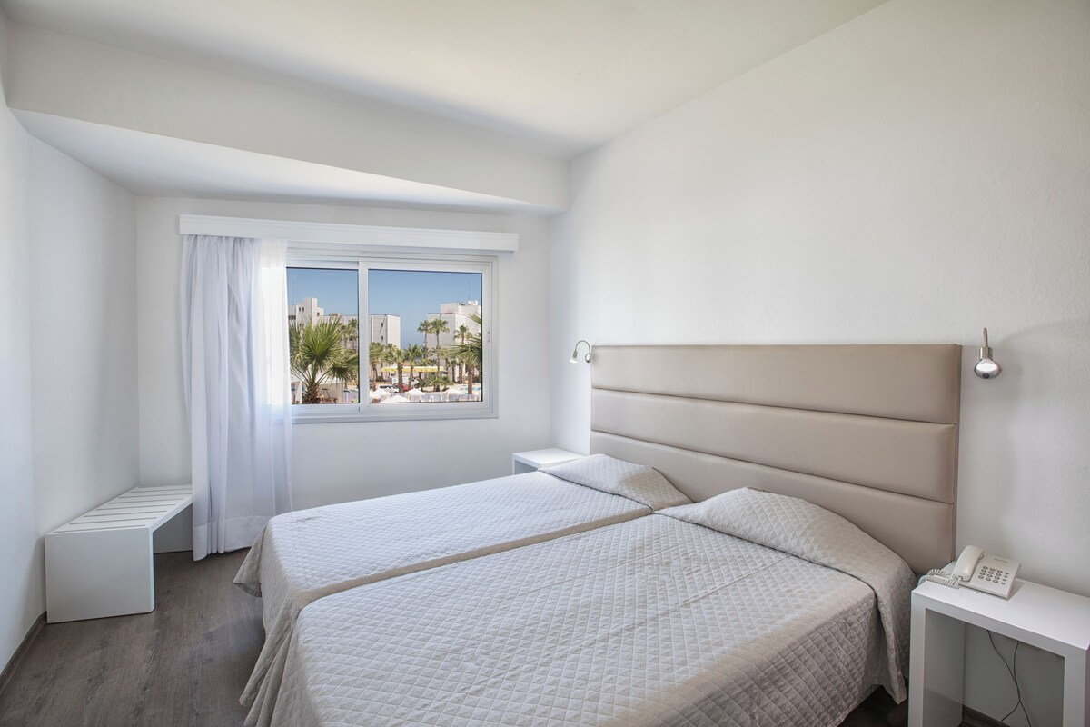 Een moderne hotelkamer met twee bedden, een groot raam en uitzicht op palmbomen en gebouwen.