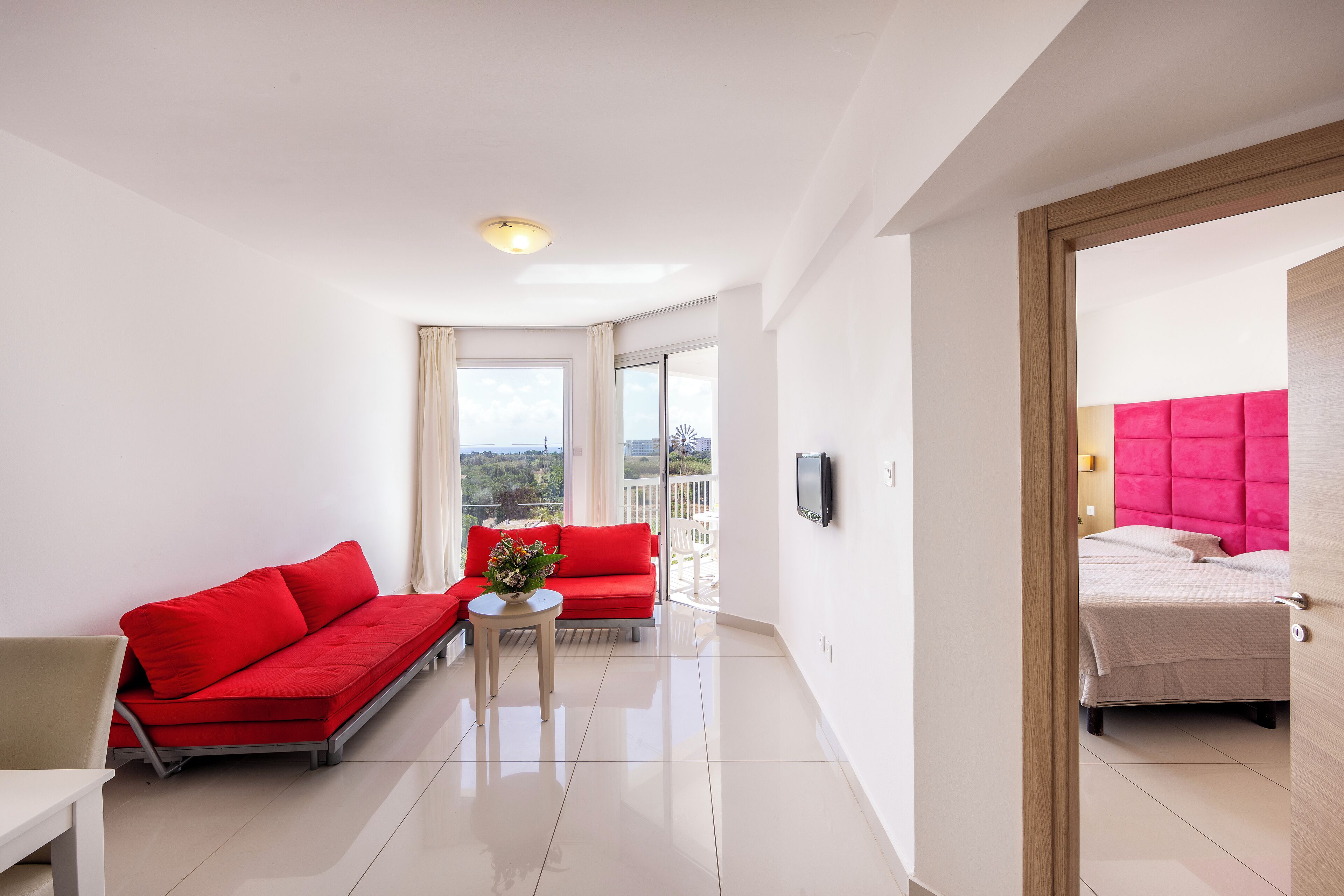 A modern hotel room with a red sofa, a small table with a vase, a bed with a pink headboard, and a balcony with a city view.