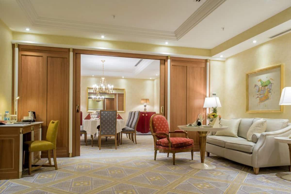 A hotel room with a dining area, a sofa, a chair, a lamp, and a painting on the wall.