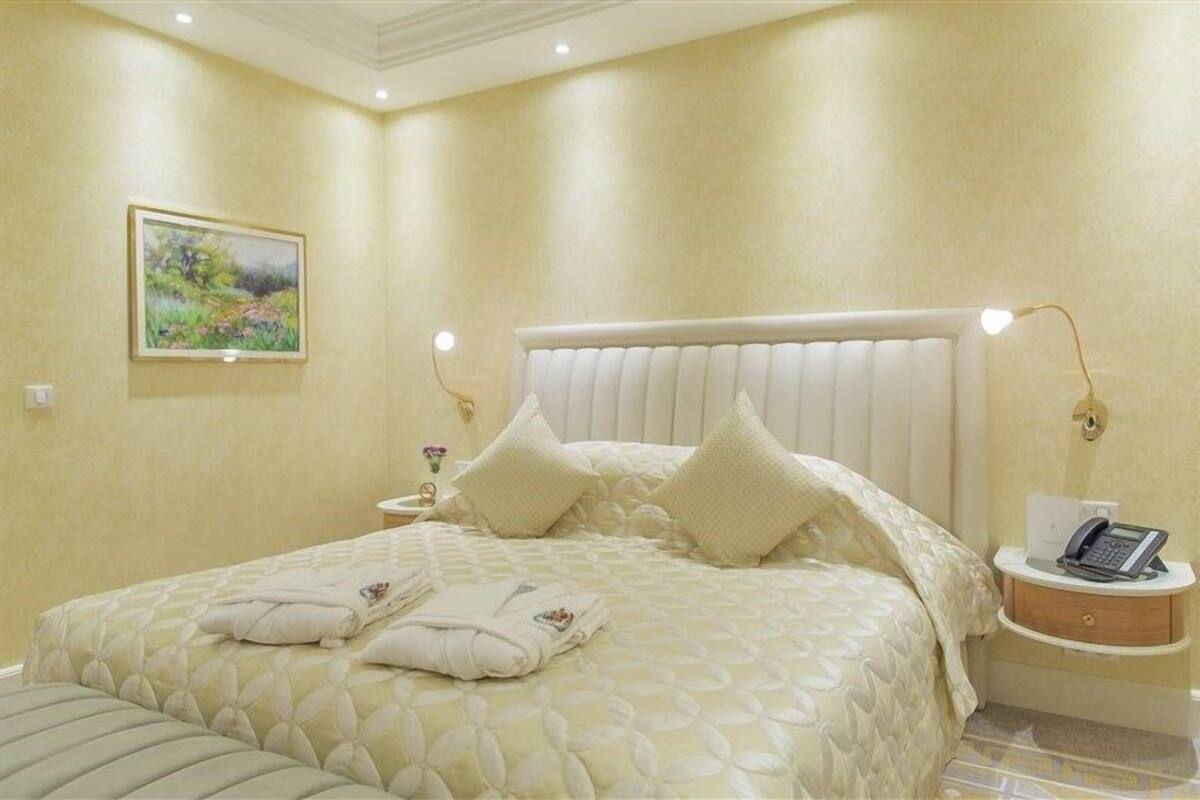 A neatly made bed with a white headboard, two bedside lamps, a bedside table with a phone, and a painting on the wall.