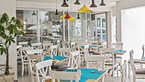 A spacious dining area with white chairs and tables, modern pendant lighting, and large windows offering views of the outside. - Hotel Napa Tsokkos (Ayia Napa)