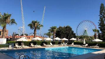 A swimming pool with lounge chairs and umbrellas, surrounded by palm trees and a Ferris wheel in the background.