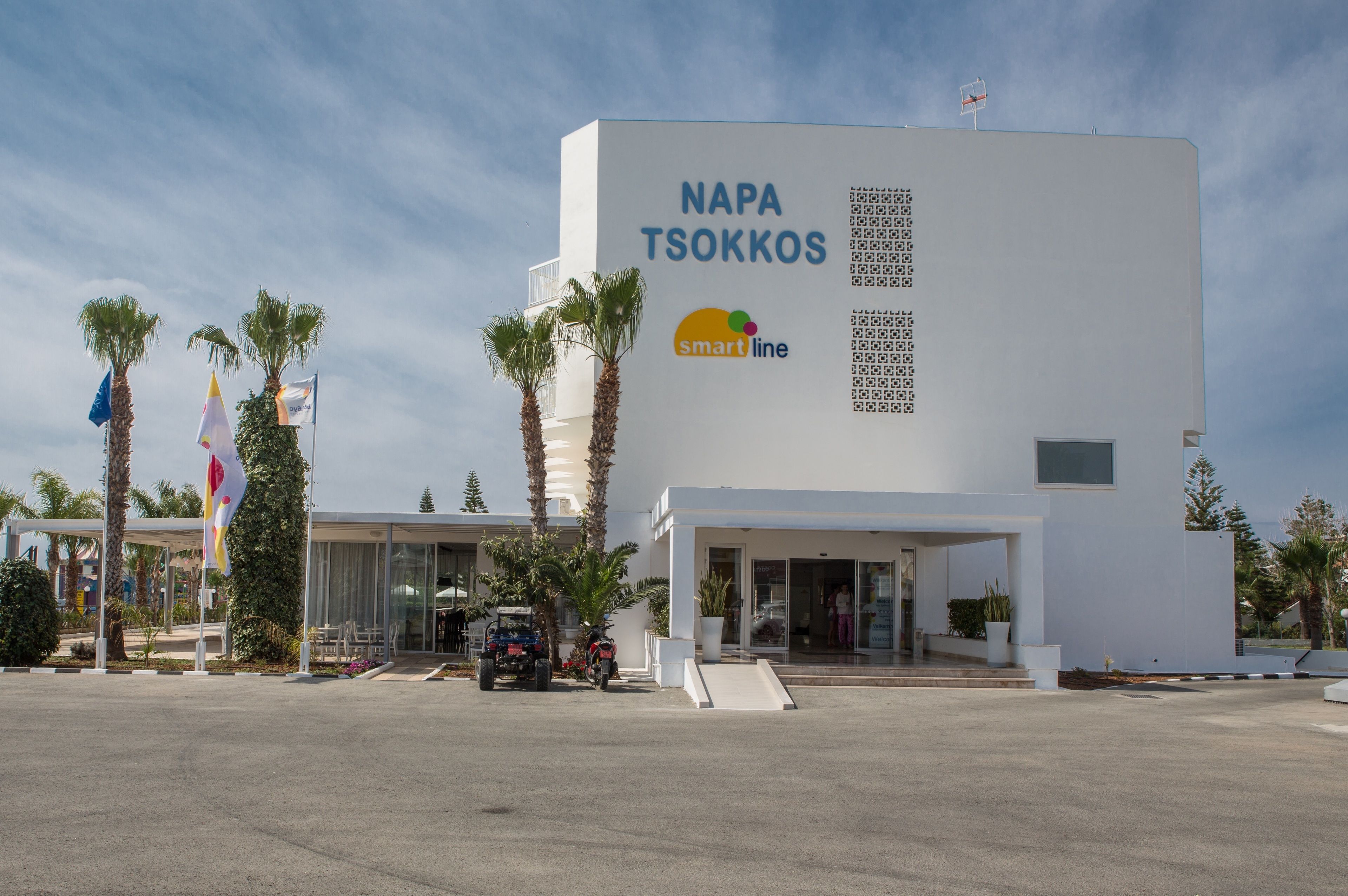 A modern building with a sign reading "NAPA TSOKKOS" and "smartline" logo, surrounded by palm trees and flags.