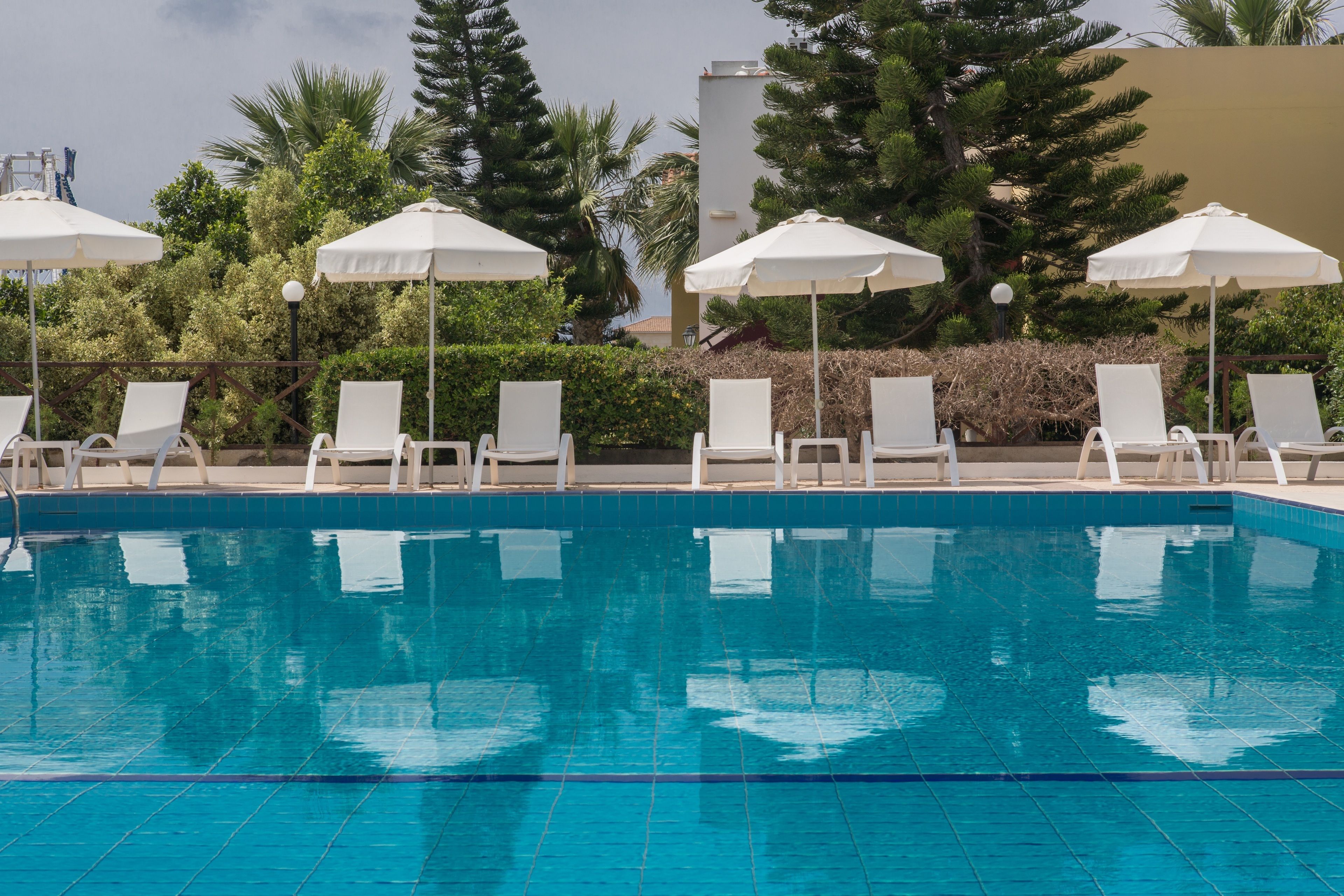 A swimming pool with white lounge chairs and umbrellas, surrounded by greenery and a building in the background.