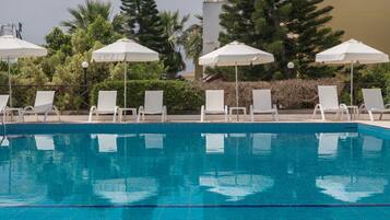 A swimming pool with white lounge chairs and umbrellas, surrounded by greenery and a building in the background.