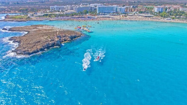 An aerial view of a coastal area with a rocky shoreline, a beach, and several boats. - Hotel Napa Tsokkos (Ayia Napa)