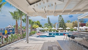 A poolside area with a bar, lounge chairs, umbrellas, and a view of the sea. - Hotel Napa Tsokkos (Ayia Napa)