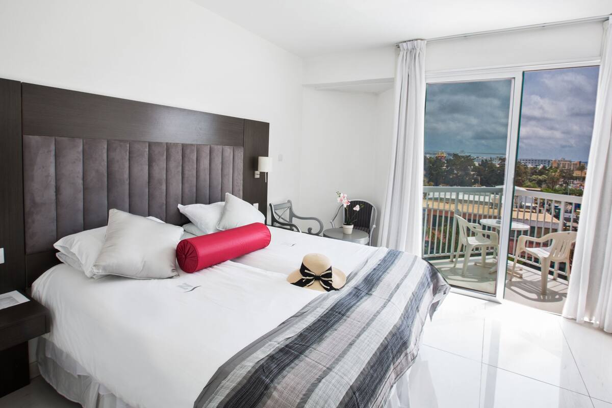 A hotel room with a large bed, a view of the city, a balcony with chairs, and a telephone on the nightstand.
