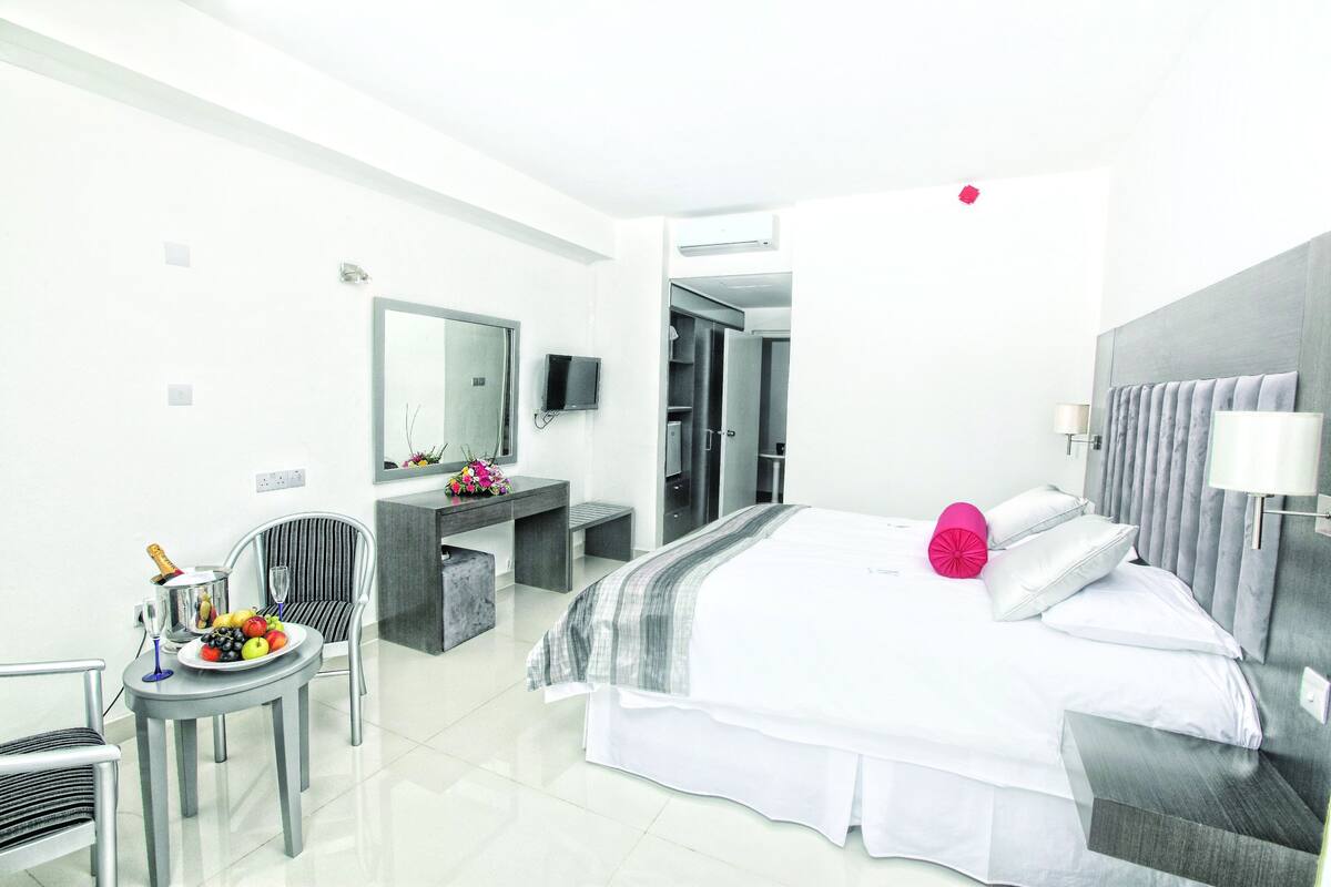 A modern hotel room with a bed, bedside table, chair, and a small desk with a mirror and TV.