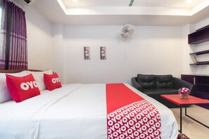 Une chambre d’hôtel avec un lit, une couverture rouge et blanche, un canapé en cuir noir, une petite table sur laquelle est posé un vase de fleurs et un ventilateur de plafond.