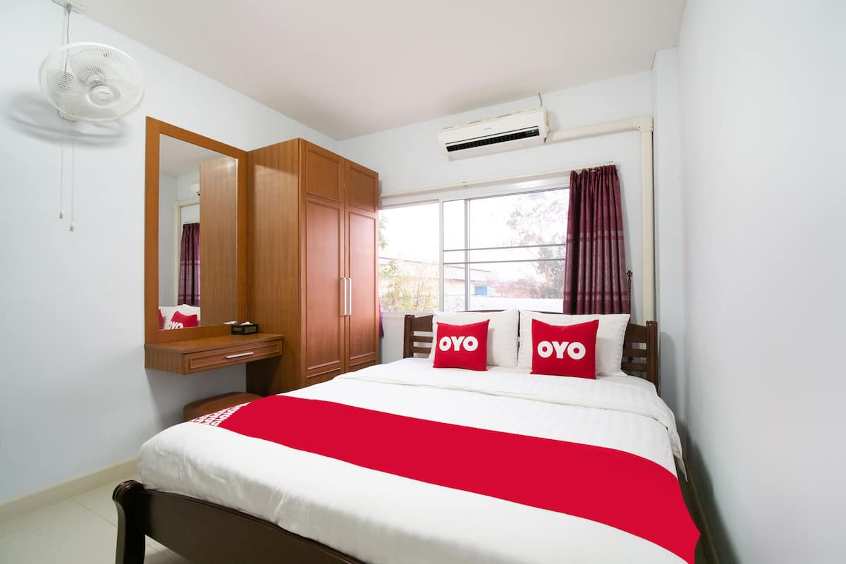 A bedroom with a bed, a wooden wardrobe, a mirror, a window with curtains, and an air conditioning unit.