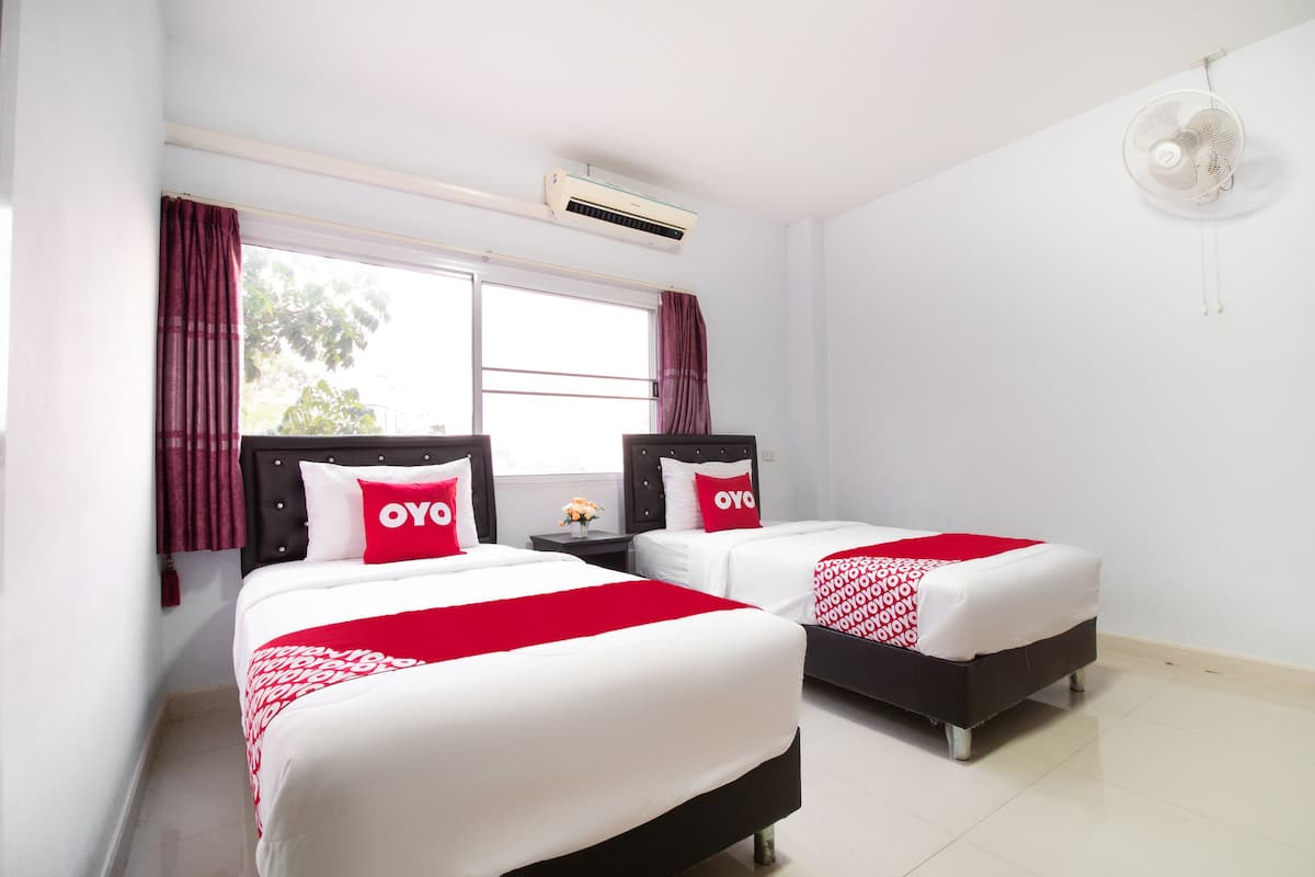 A hotel room with two beds, each with a red and white bedspread and matching pillows. There is a window with curtains, an air conditioning unit, and a ceiling fan.