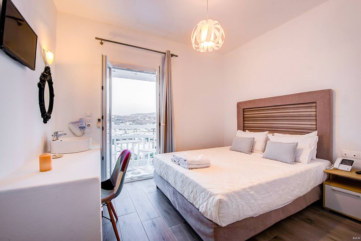 A hotel room with a large bed, a bedside table with a phone, a chair, a window with curtains, a wall-mounted TV, and a bathroom with a sink and mirror.