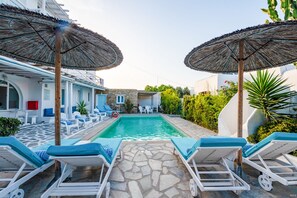 Ένας χώρος δίπλα στην πισίνα με ξαπλώστρες και ομπρέλες, ένα δάπεδο επενδεδυμένο με πέτρα και μια κρυστάλλινη, γαλάζια πισίνα.