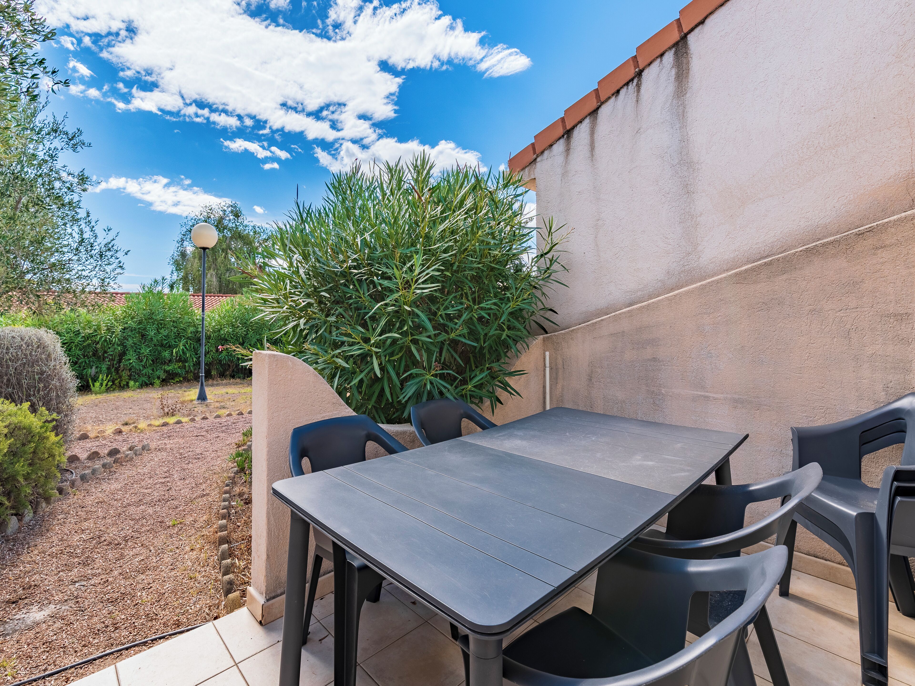 Un espace extérieur aménagé en terrasse, avec une table et des chaises, entouré de plantes et surplombé d’un ciel bleu clair.