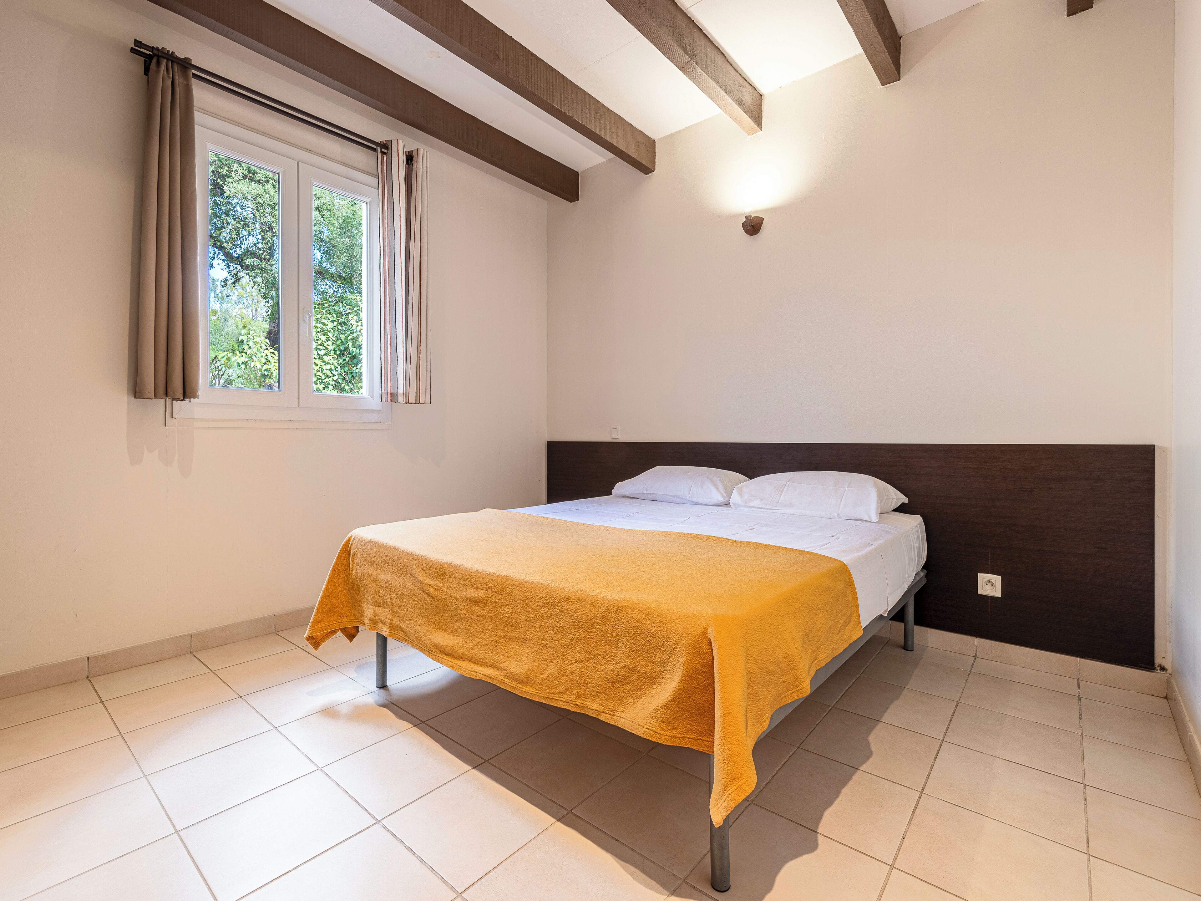 Een slaapkamer met een bed, een raam met gordijnen, een houten balk in het plafond en een wandlamp.