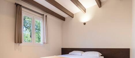 Een slaapkamer met een bed, een raam met gordijnen, een houten balk in het plafond en een wandlamp.