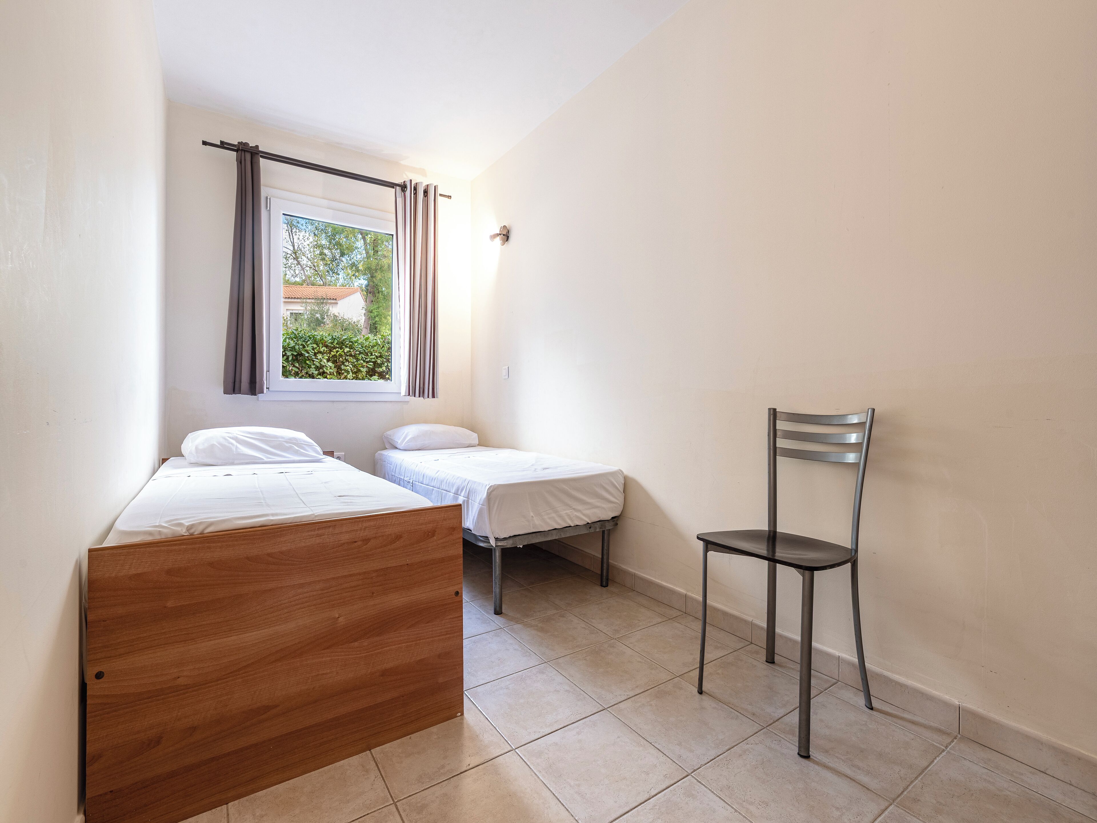 Une petite pièce propre, avec deux lits, une chaise et une fenêtre avec des rideaux.