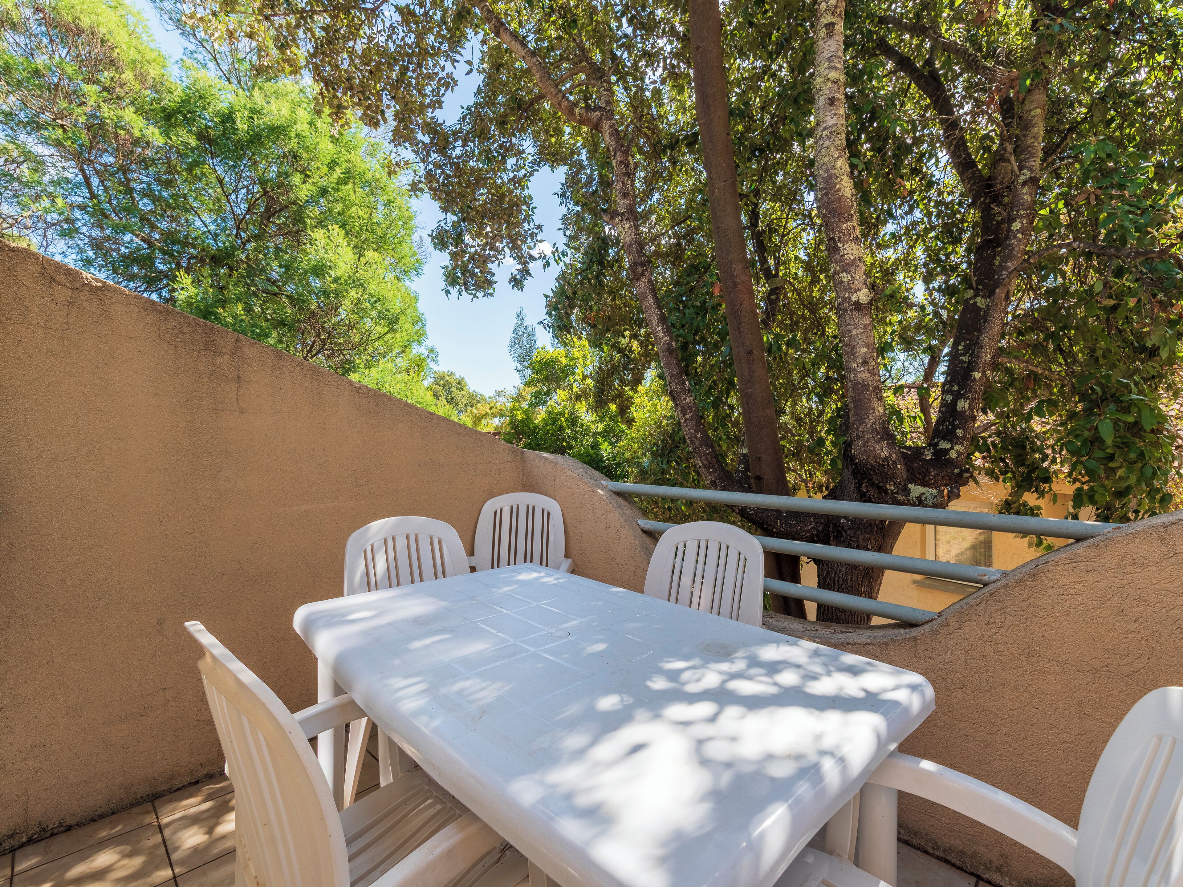 An outdoor patio with a table and chairs, surrounded by trees and a beige wall.
