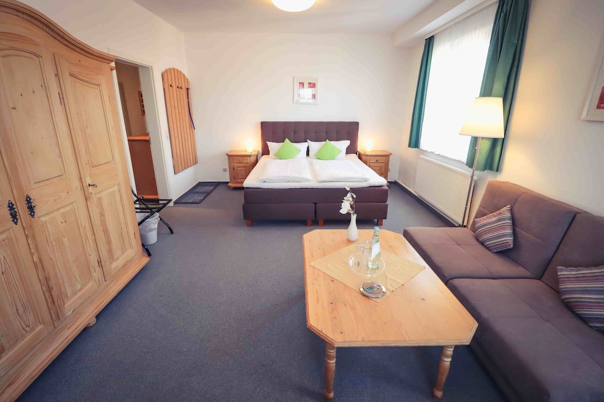 Habitación de hotel con cama, sofá, mesa de madera, armario y ventana con cortinas.