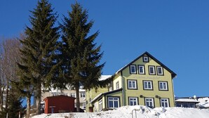 Un edificio de varios pisos con fachada verde y numerosas ventanas, rodeado de nieve y árboles altos.