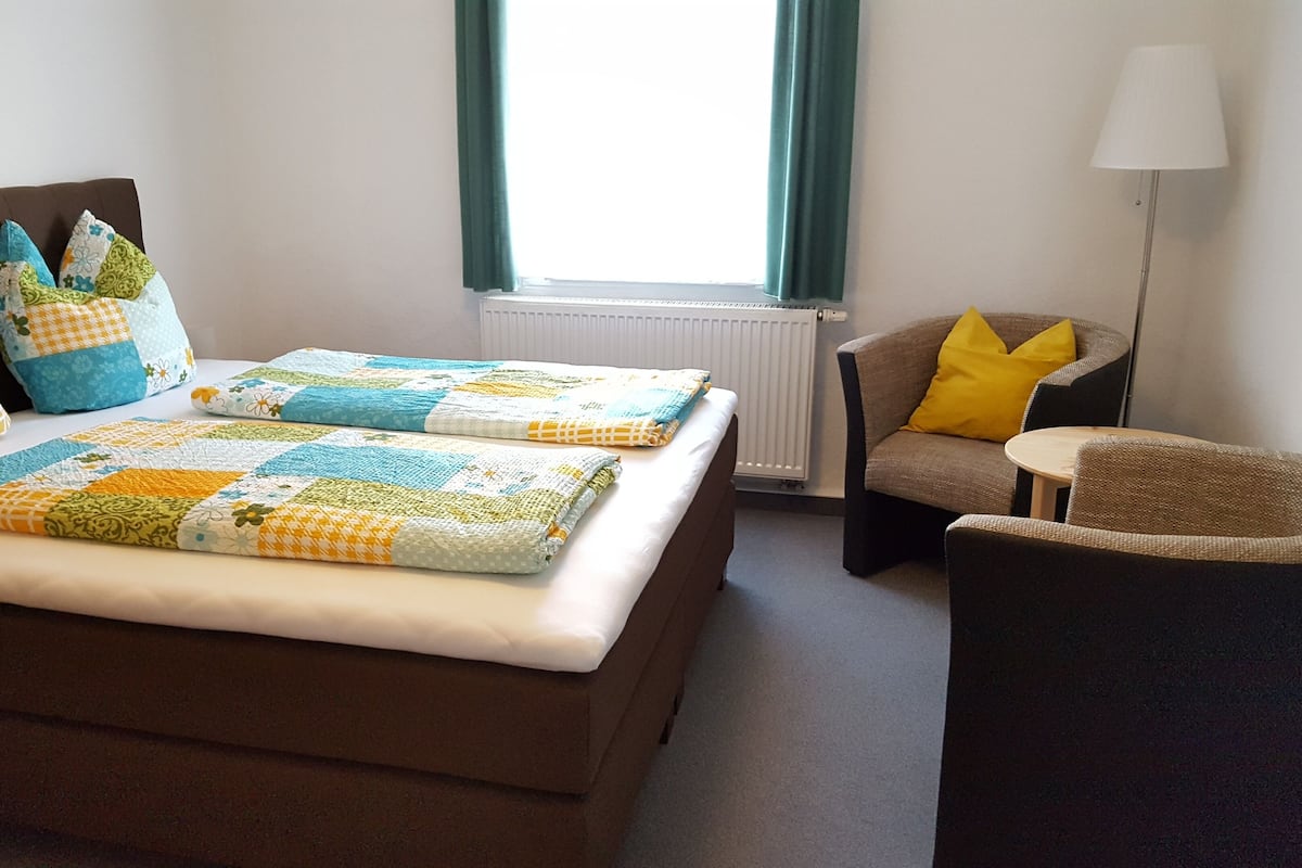 Habitación de hotel con cama, dos almohadas, una silla, una lámpara y una ventana con cortinas.