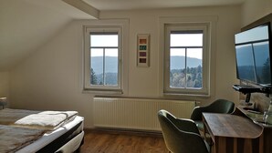Un dormitorio con cama, escritorio, dos sillas, televisor y dos ventanas con vistas a las montañas.