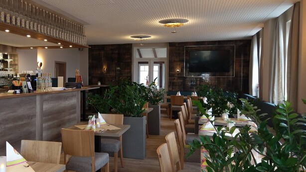 Un restaurante con mesas y sillas de madera, plantas en macetas y una zona de bar con botellas.