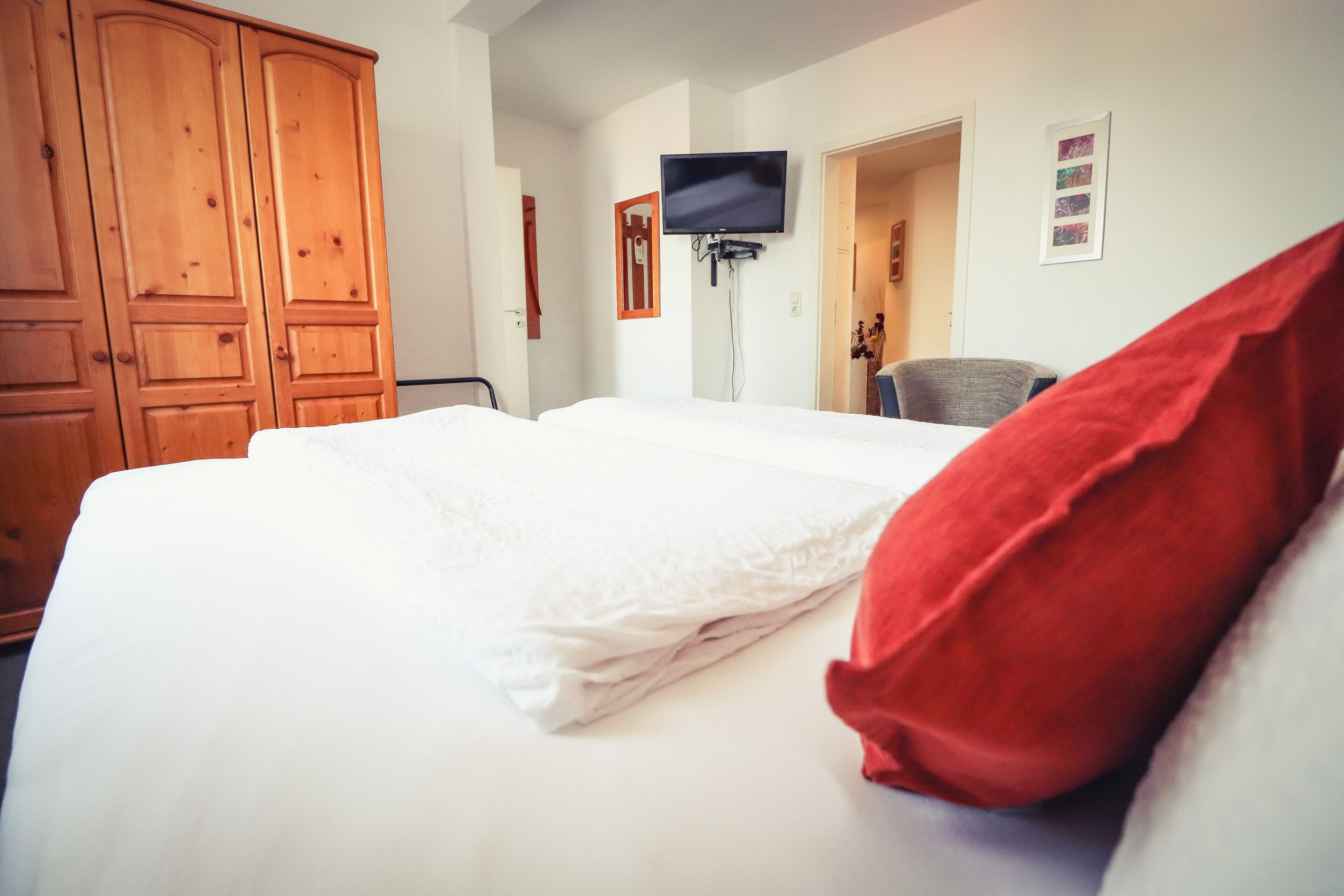 A hotel room with a large bed, a television mounted on the wall, wooden wardrobe, and a red pillow.