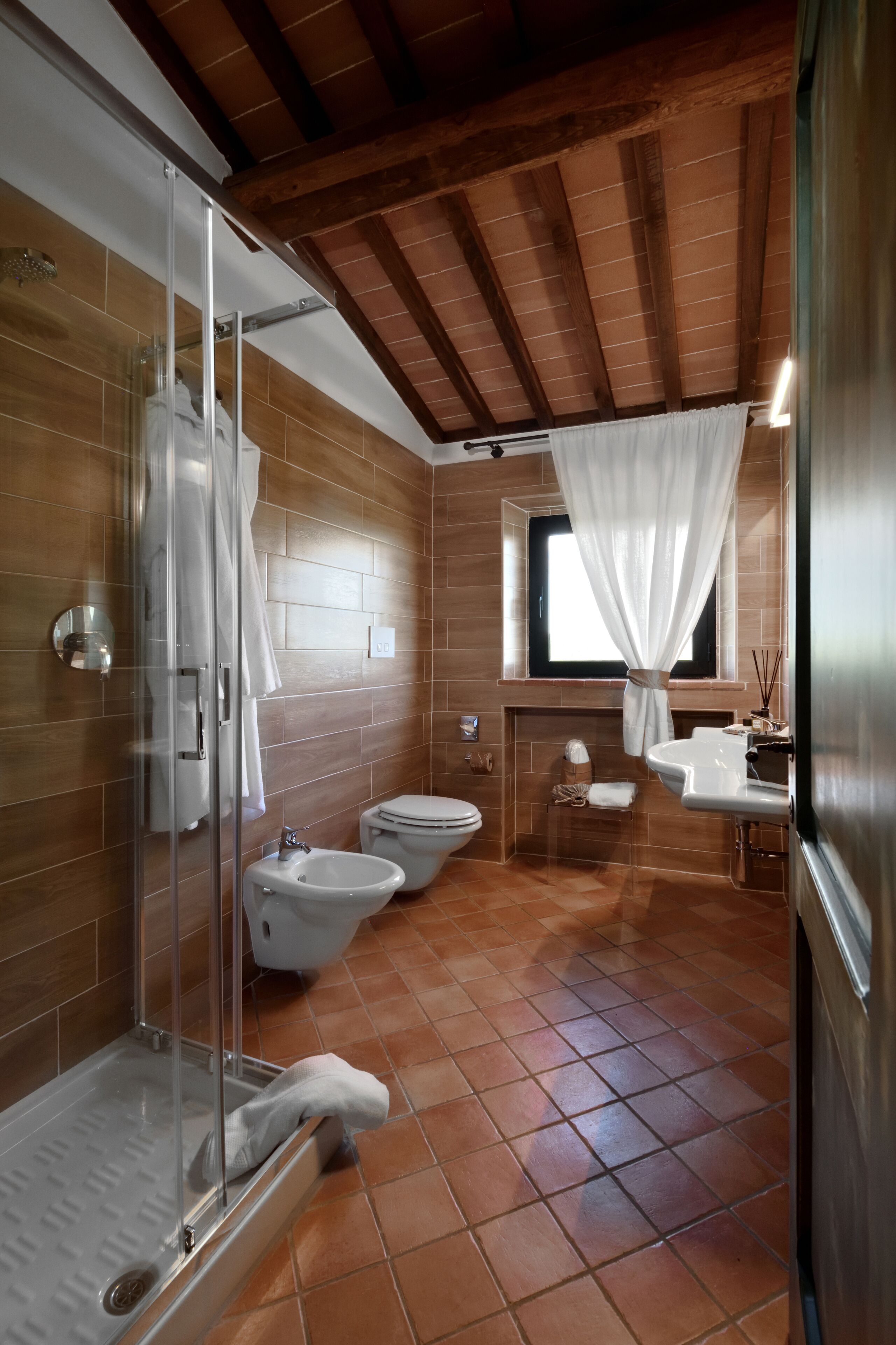 A bathroom with a glass shower enclosure, two toilets, a sink, and a window with a curtain.