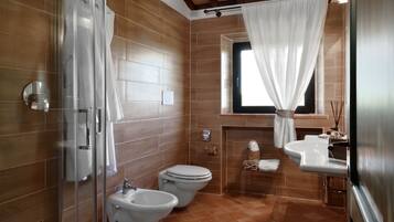 A bathroom with a glass shower enclosure, two toilets, a sink, and a window with a curtain.