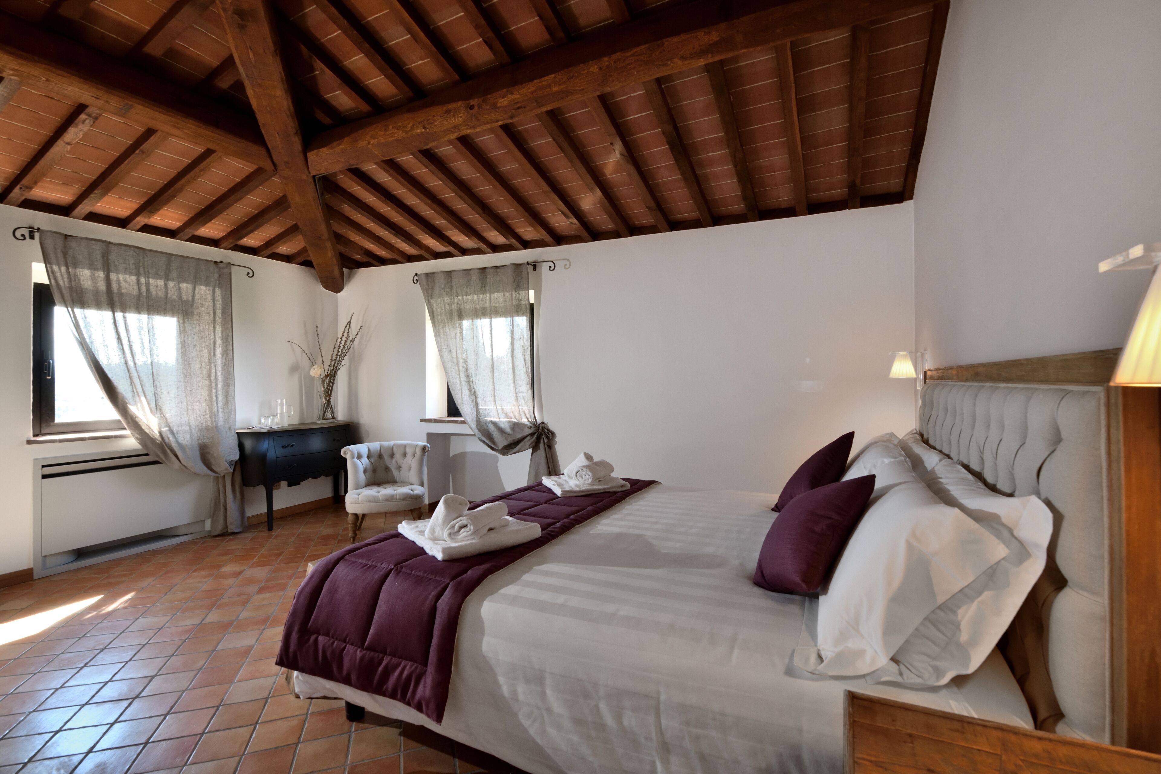 A bedroom with a large bed, a desk, and a chair. There are curtains on the windows, and the room has a wooden ceiling.