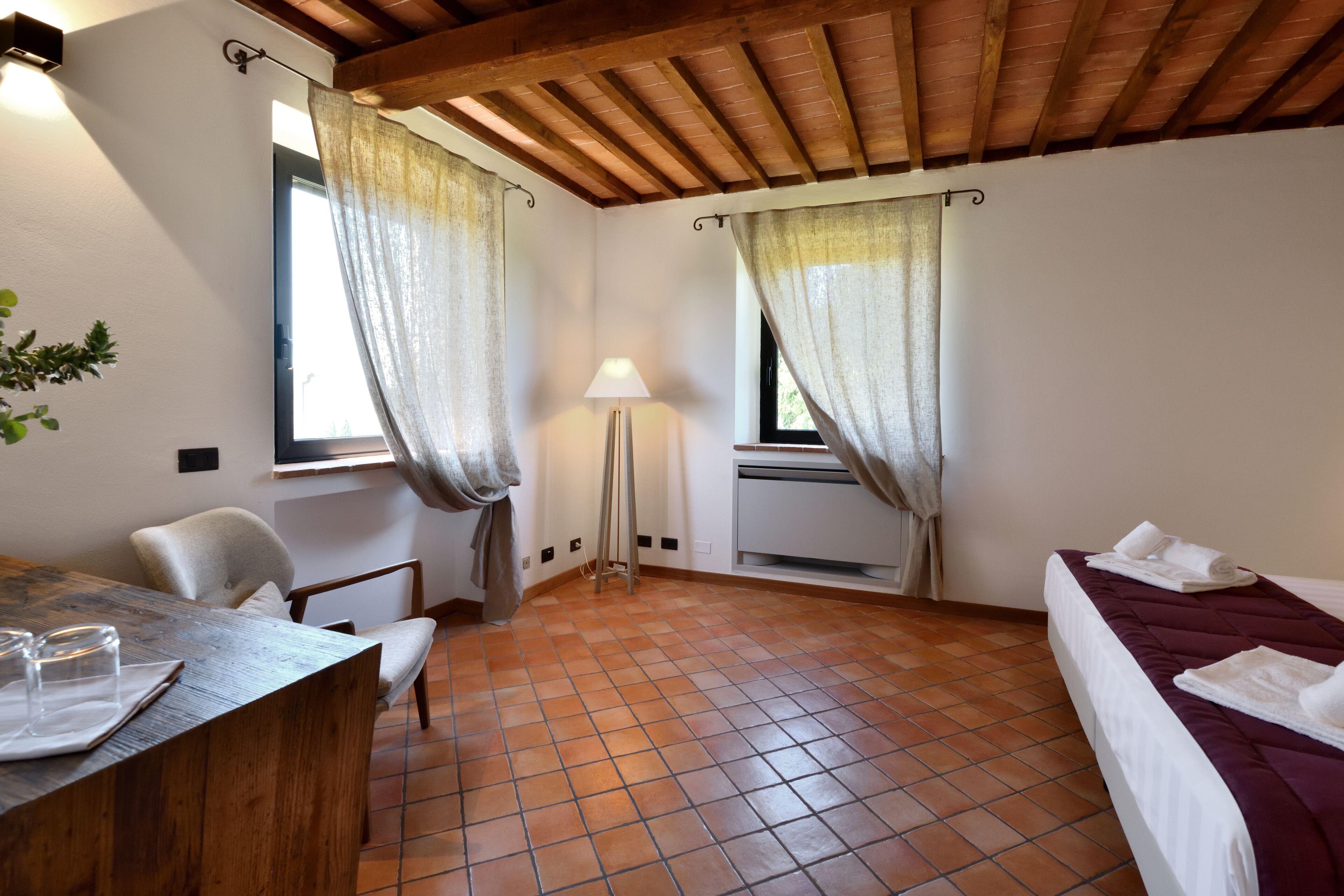 Quarto com teto de madeira, piso de azulejos, cama com roupa de cama branca, escrivaninha com cadeira e janela com cortinas.