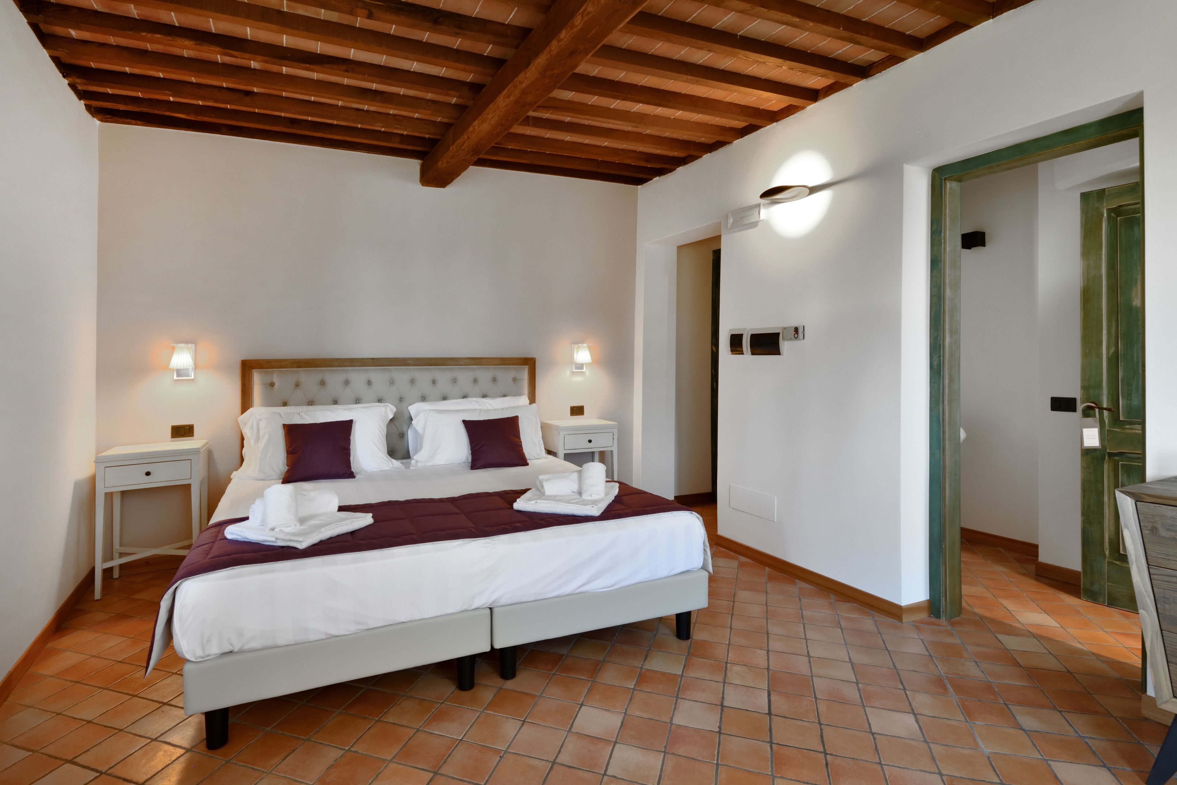 Una cama bien hecha con sábanas blancas y un edredón color burdeos, dos mesitas de noche con lámparas y un techo de madera.