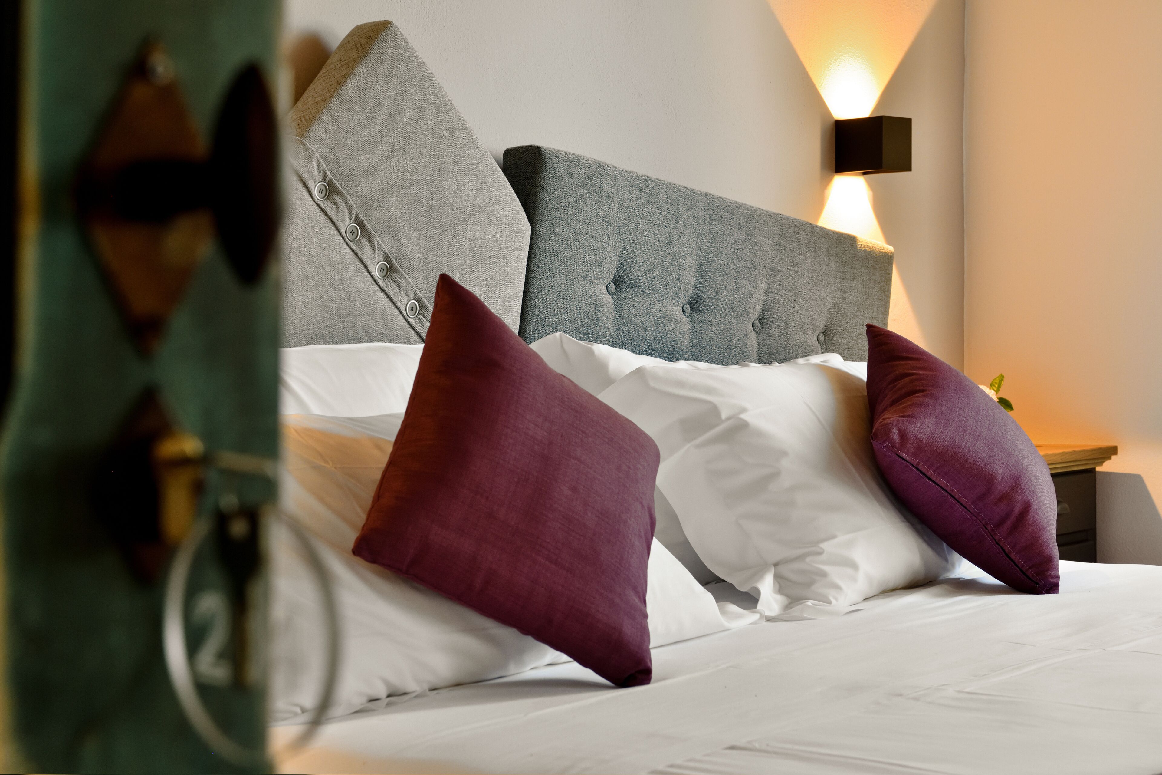 Un lit bien fait, avec du linge de lit blanc et deux coussins violets, une tête de lit rembourrée grise et un luminaire fixé au mur.