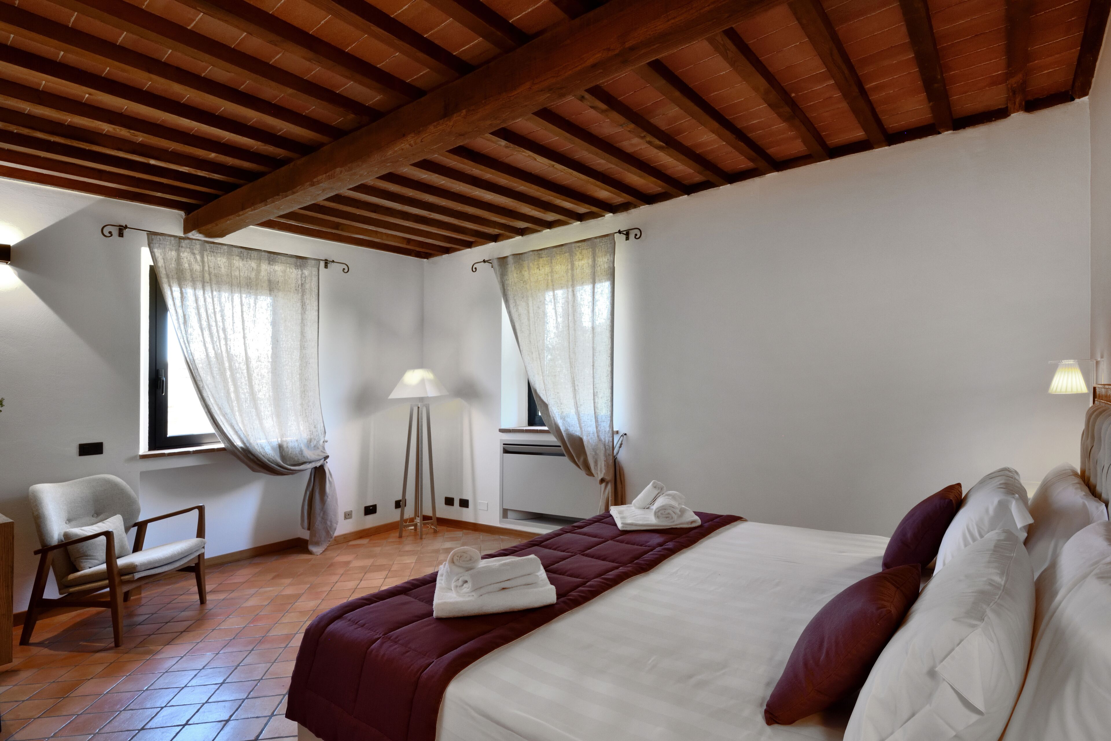 Un dormitorio con techo de madera, una cama con ropa de cama blanca y granate, una silla y dos ventanas con cortinas de gasa.