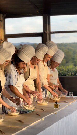 Un groupe de chefs, coiffés de leurs toques blanches et portant des tabliers, prépare des plats autour d’une longue table, avec une vue imprenable sur un paysage montagneux.