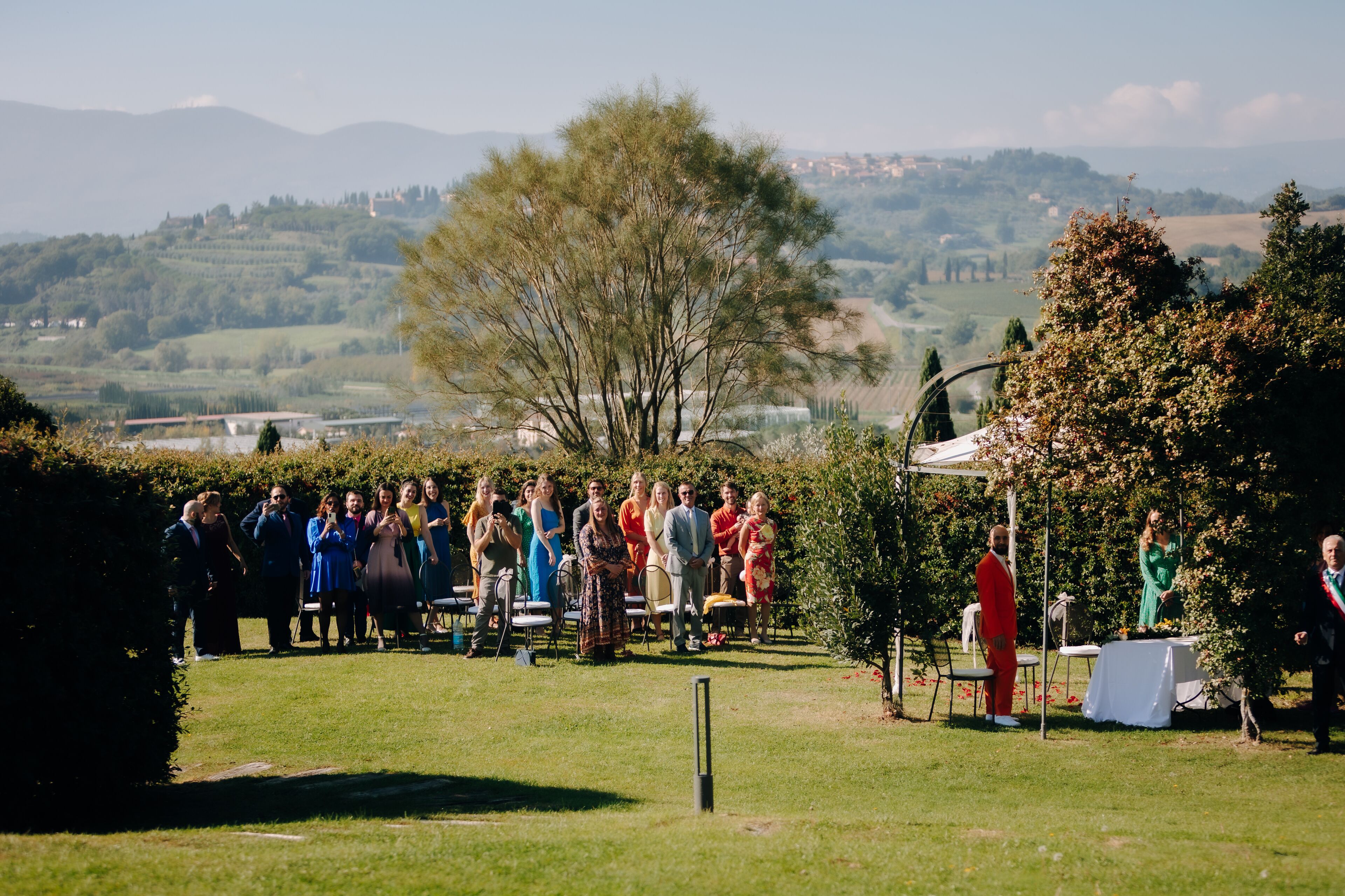 A group of people gathered outdoors, possibly at a wedding or event, with a scenic view of hills and a clear sky in the background.