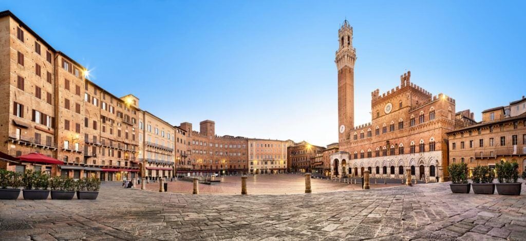 A historic square with a prominent tower, stone buildings, and potted plants.