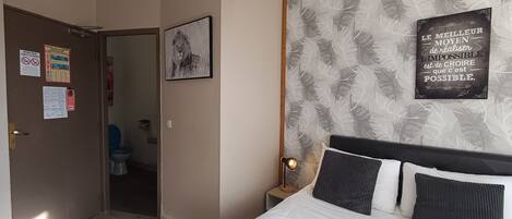 Un dormitorio con cama, una mesita de noche con lámpara, un cartel en la pared y una puerta que da al baño.
