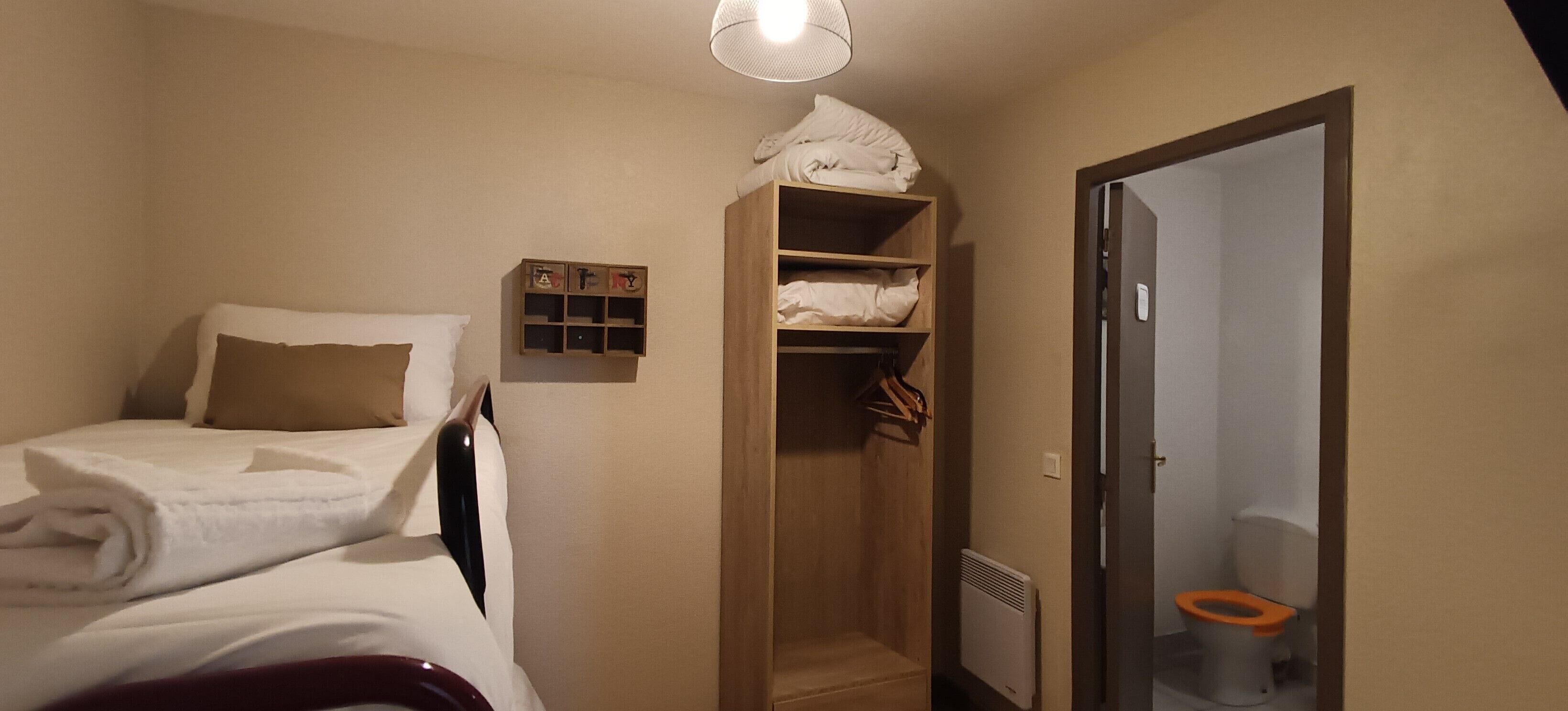 A small, single-bed room with a wooden wardrobe, a small shelf with letters, and a toilet with an orange seat.