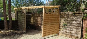 Una estructura de madera con techo de listones y un portón, rodeada por una cerca de madera y árboles.