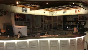 Una zona de bar con un mostrador blanco curvo, estantes con botellas y vasos, una computadora ASUS y un techo con vigas a la vista.