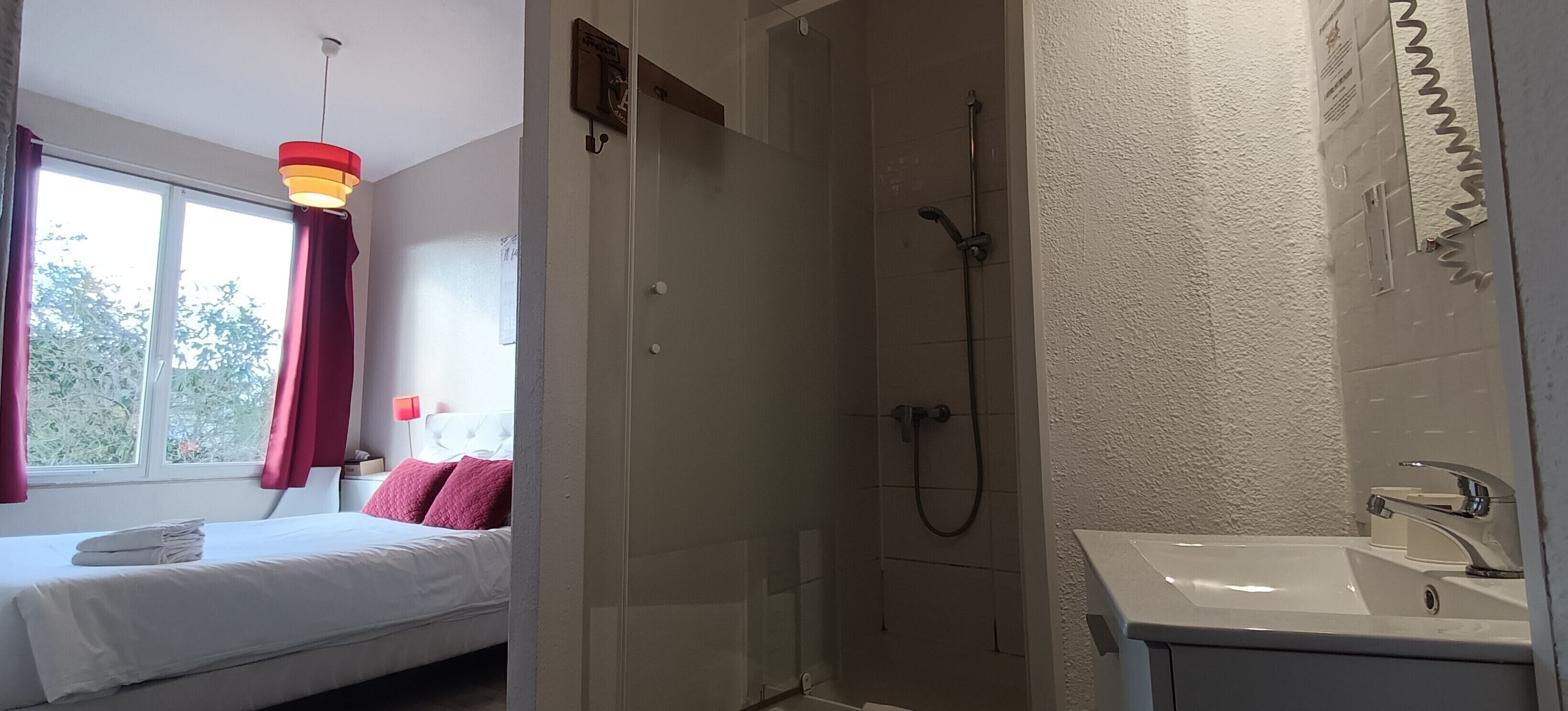 A hotel room with a bed, a window with curtains, a shower area, and a sink.