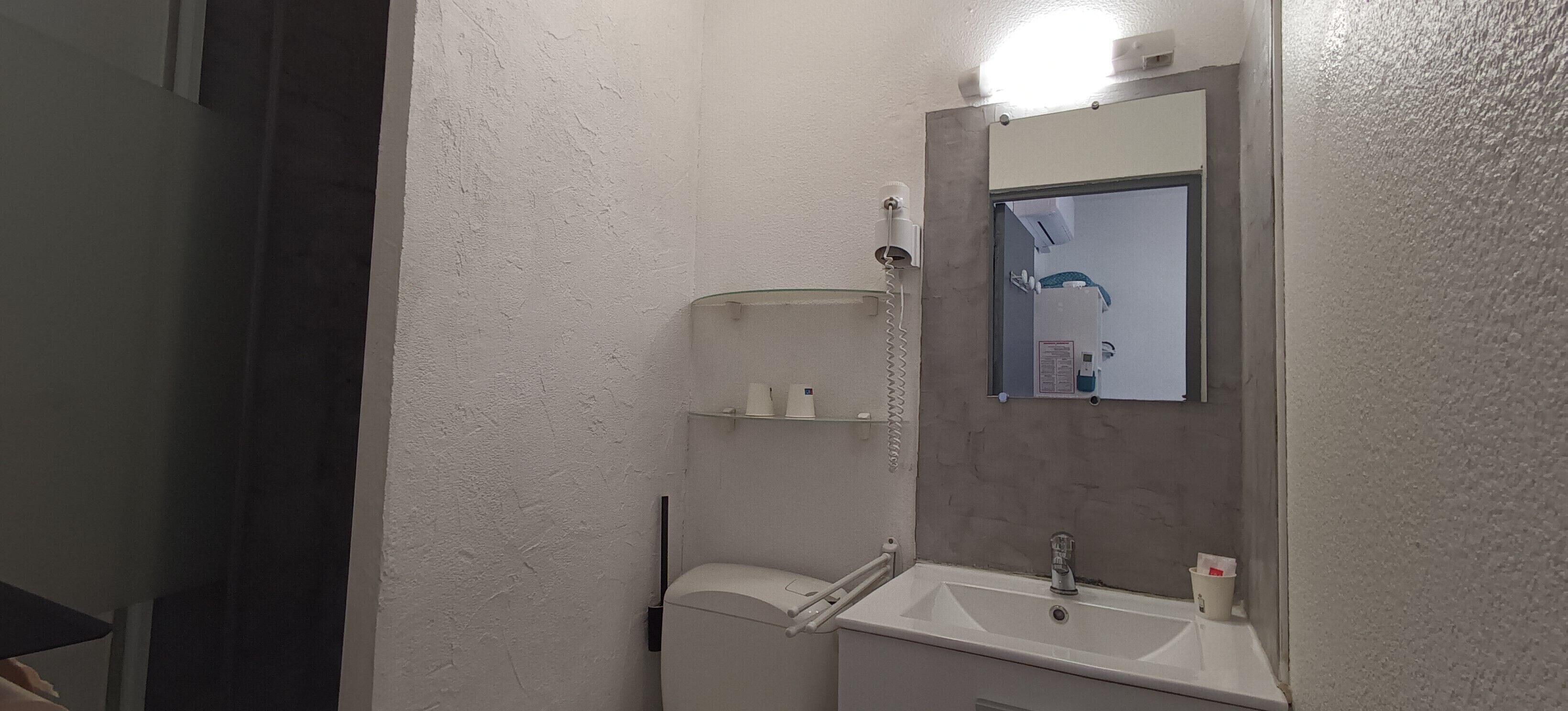 Un baño pequeño con un lavamanos blanco, espejo y un inodoro. Hay repisas sobre el lavamanos y un perchero en la pared.