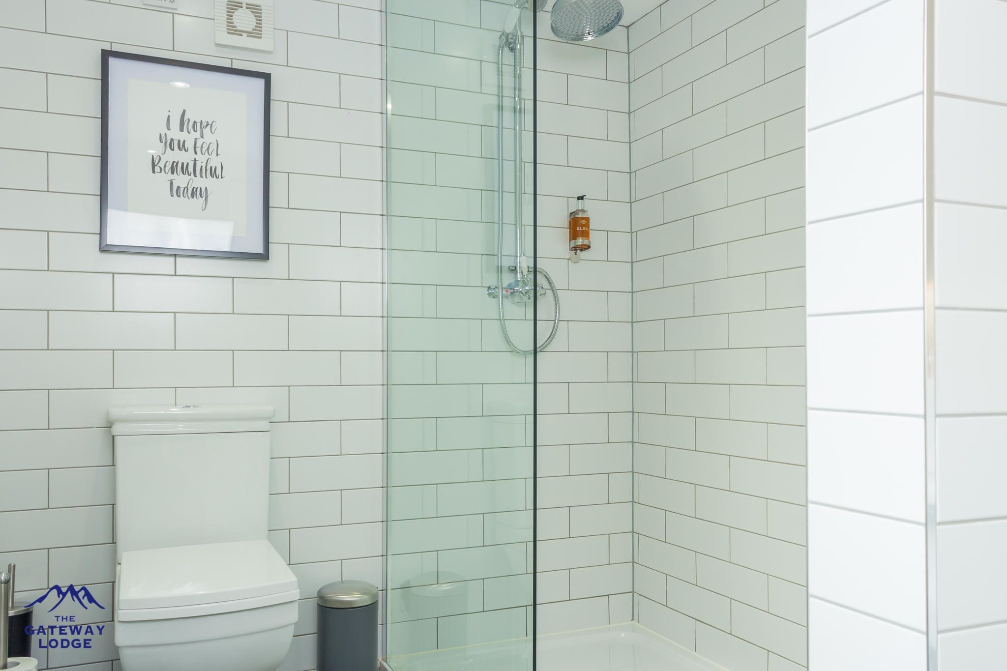 A modern bathroom with white tiles, a glass shower enclosure, a framed inspirational quote, and a toilet.