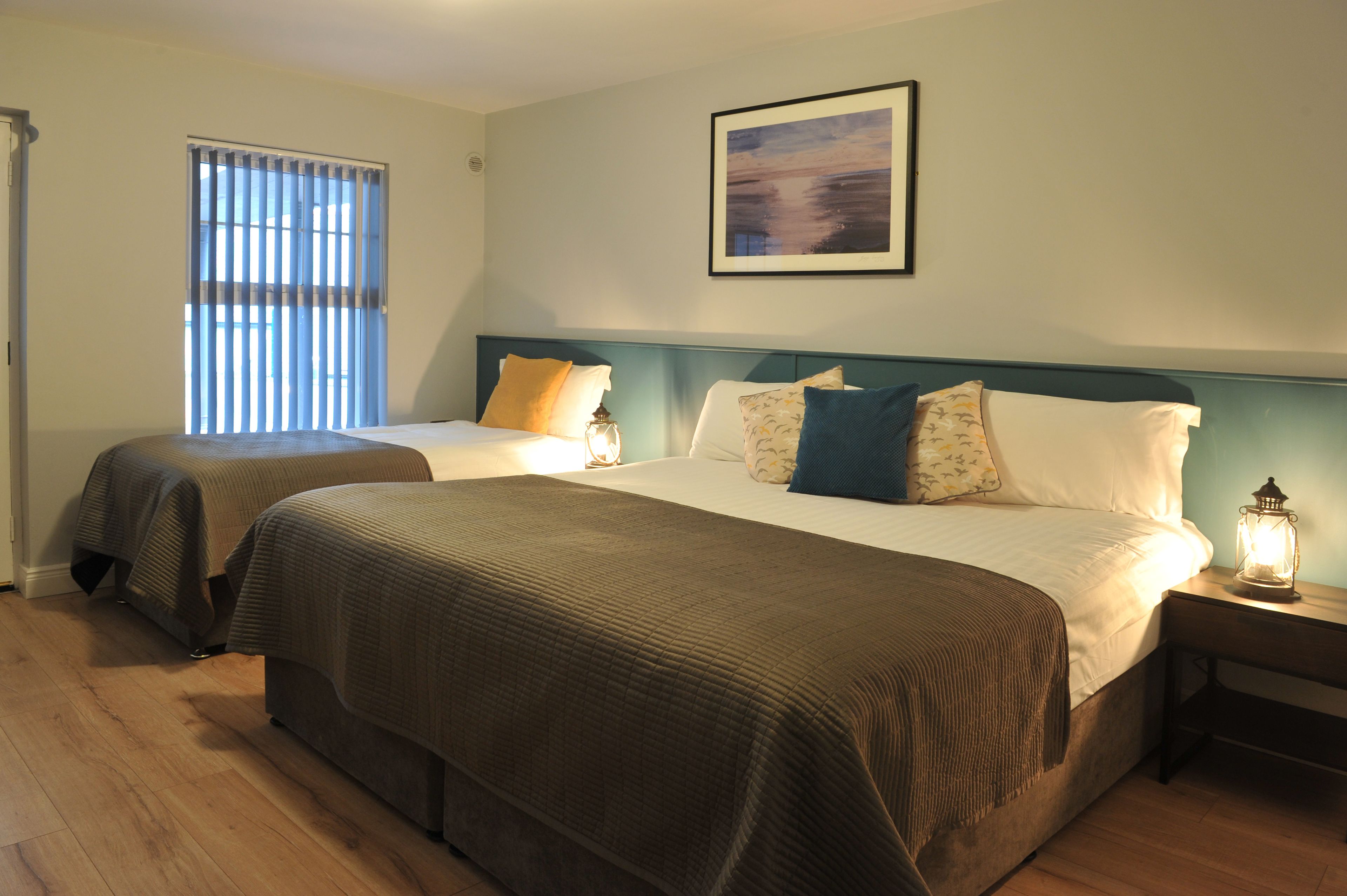 A hotel room with two beds, a window with blinds, a framed picture on the wall, and a nightstand with a lantern.