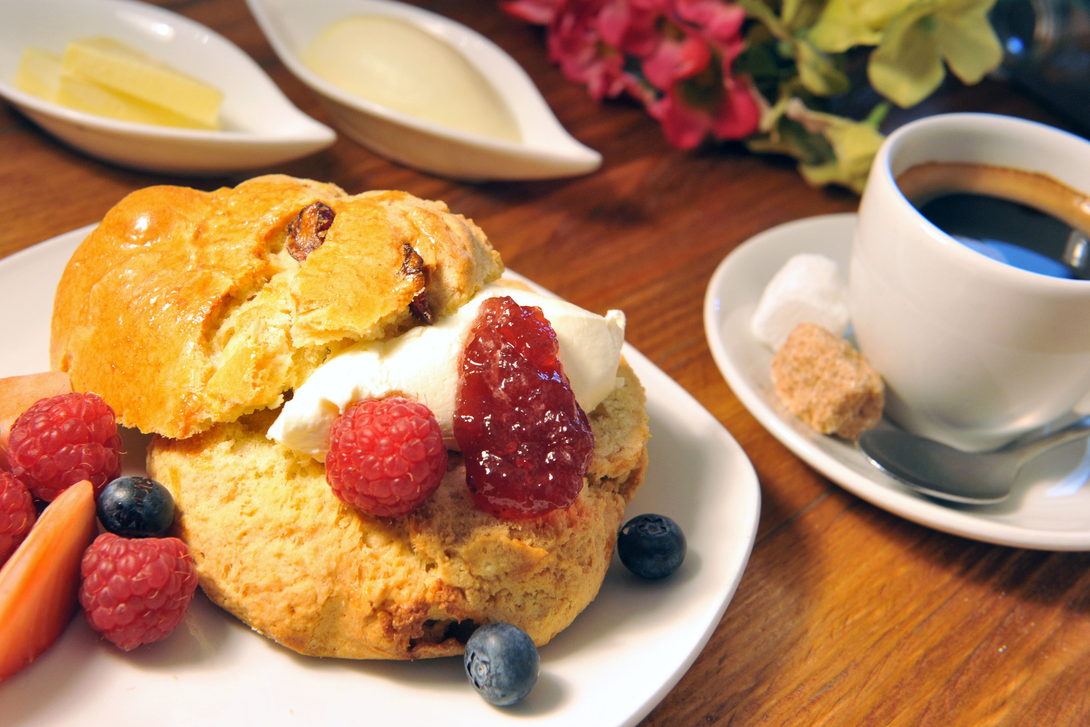 A plate with a scone, raspberries, and blueberries, accompanied by a cup of coffee and a small dish of butter.