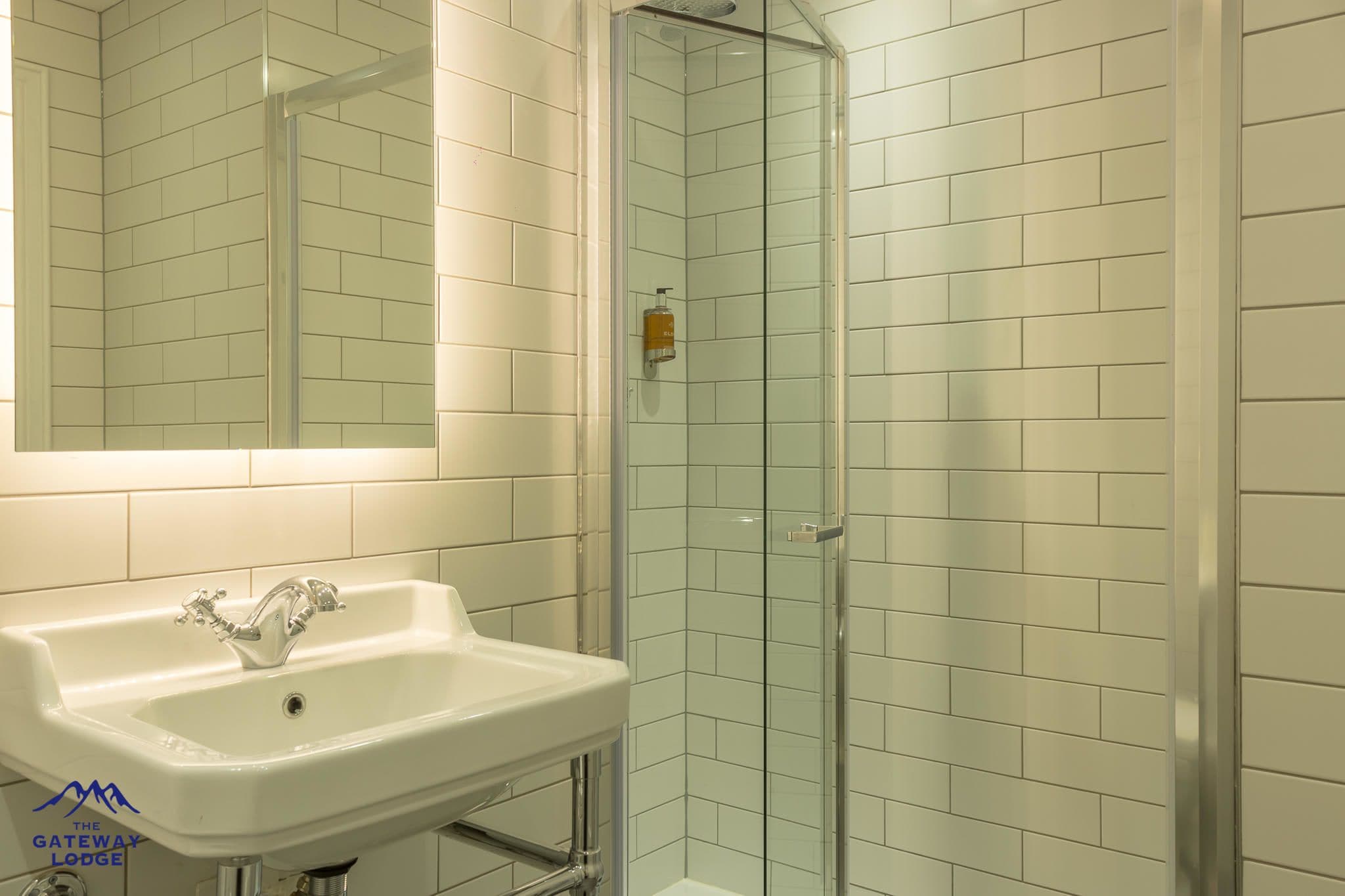 Une salle de bain avec un lavabo blanc, une cabine de douche en verre et des murs recouverts de carreaux blancs.